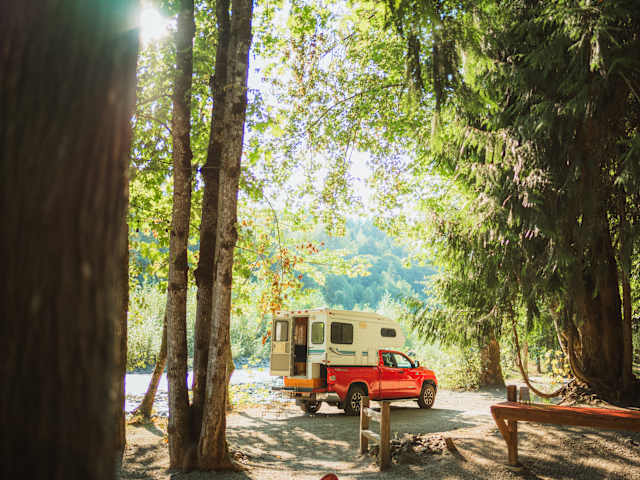 Red truck camper parked in dappled forest clearing near river with picnic table