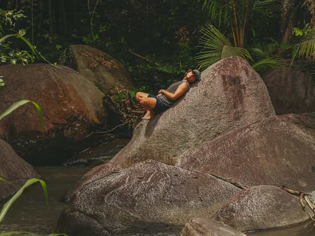 Person resting on large river boulder surrounded by dense tropical forest