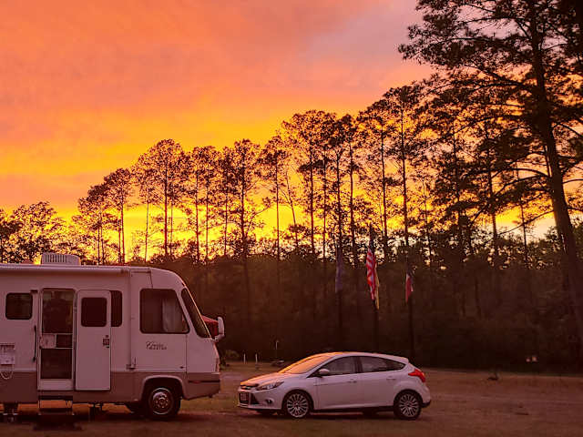 RV and car parked under a fiery orange sunset with silhouetted pine trees