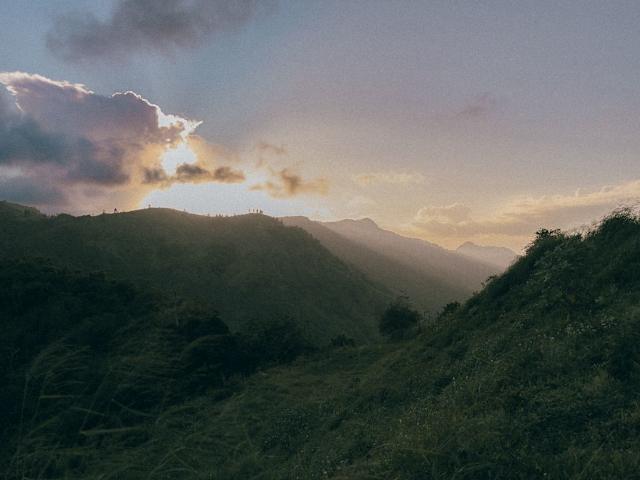 Sunset breaking through clouds over layered mountain ridges with silhouetted hikers on a distant ridge