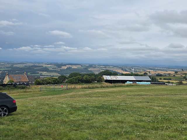 Rolling green farmland with barn and hedgerows stretching toward distant hills under cloudy sky